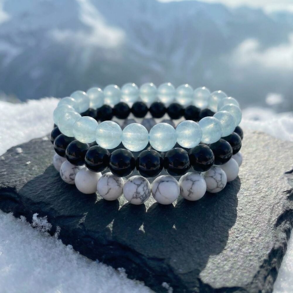 Stacked Glacial Calm Bracelet Triad on charcoal stone surrounded by winter snow — Obsidian, Aquamarine and Howlite beads in natural light.