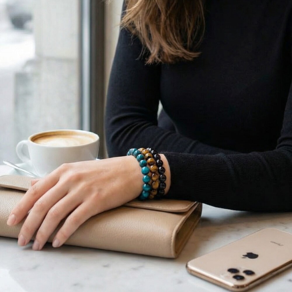 Woman’s hand resting on a beige clutch bag on a polished white-grey marble café table in Zurich, wearing the Alpine Courage bracelet triad — a grounded, elegant moment of calm confidence.