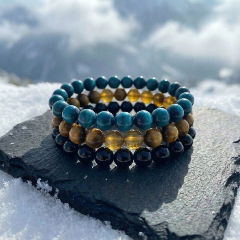 The Alpine Courage bracelet triad stacked together on a charcoal rock surrounded by snow — rich golden, deep blue, and matte black stones reflecting the strength and clarity of high winter ascents.