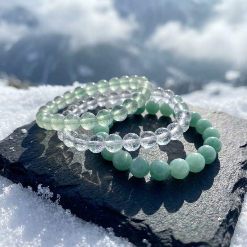 The Alpine Renewal bracelet triad in a cascade arrangement on a charcoal rock surrounded by snow, with a soft-focus meltwater Alpine backdrop — pale green fluorite, mint amazonite, and ice quartz catching the winter light