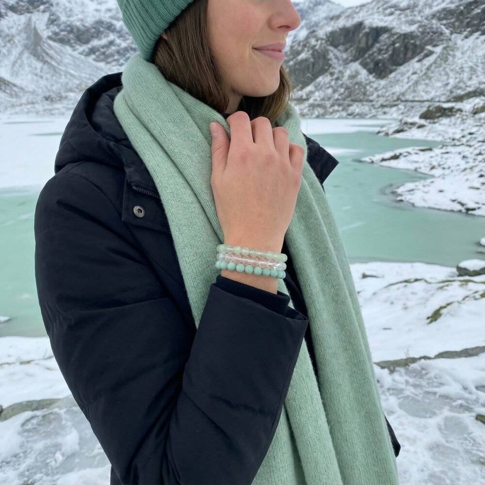 Woman overlooking pale green frozen Alpine lakes, gently adjusting her pale mint scarf, wearing the Alpine Renewal bracelet triad — a quiet moment of clarity, freshness, and emotional reset.