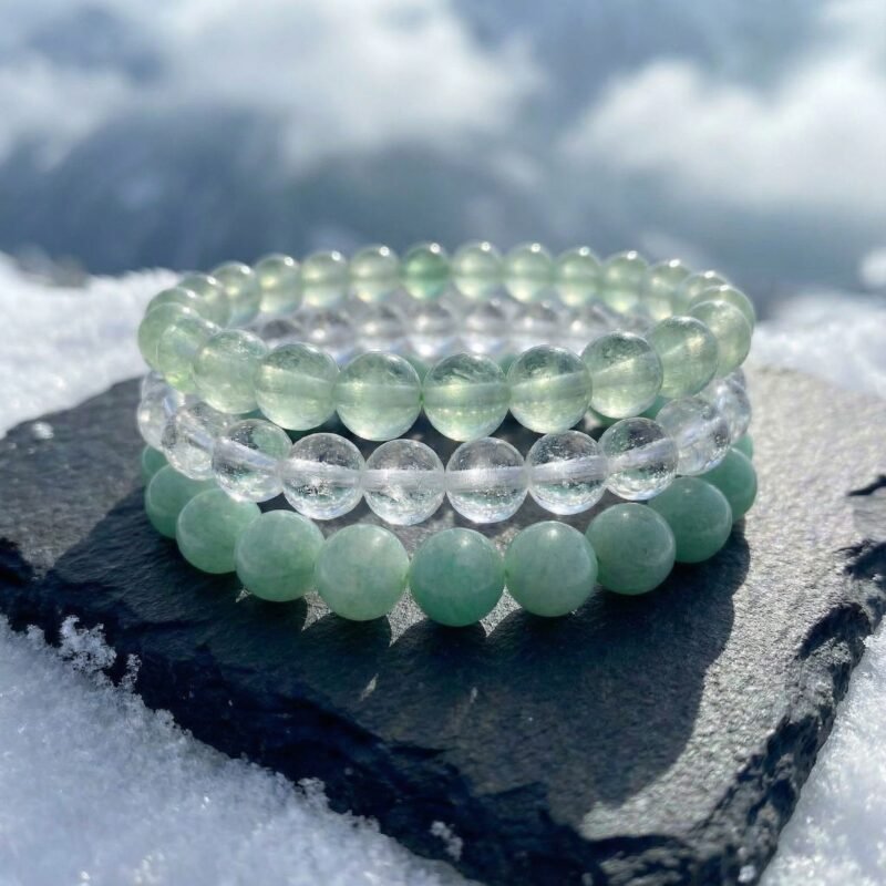 The Alpine Renewal bracelet triad stacked together on a charcoal rock surrounded by snow, showcasing pale green fluorite, mint amazonite, and clear ice quartz in a bright, renewing Alpine palette.