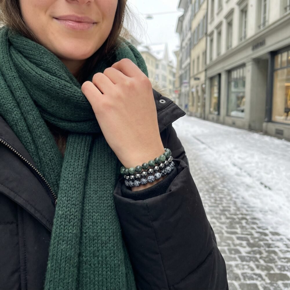Woman walking down a snowy Zurich backstreet, adjusting a dark green woollen scarf, wearing the Winter Resilience bracelet triad — a quiet moment of grounded strength and endurance.