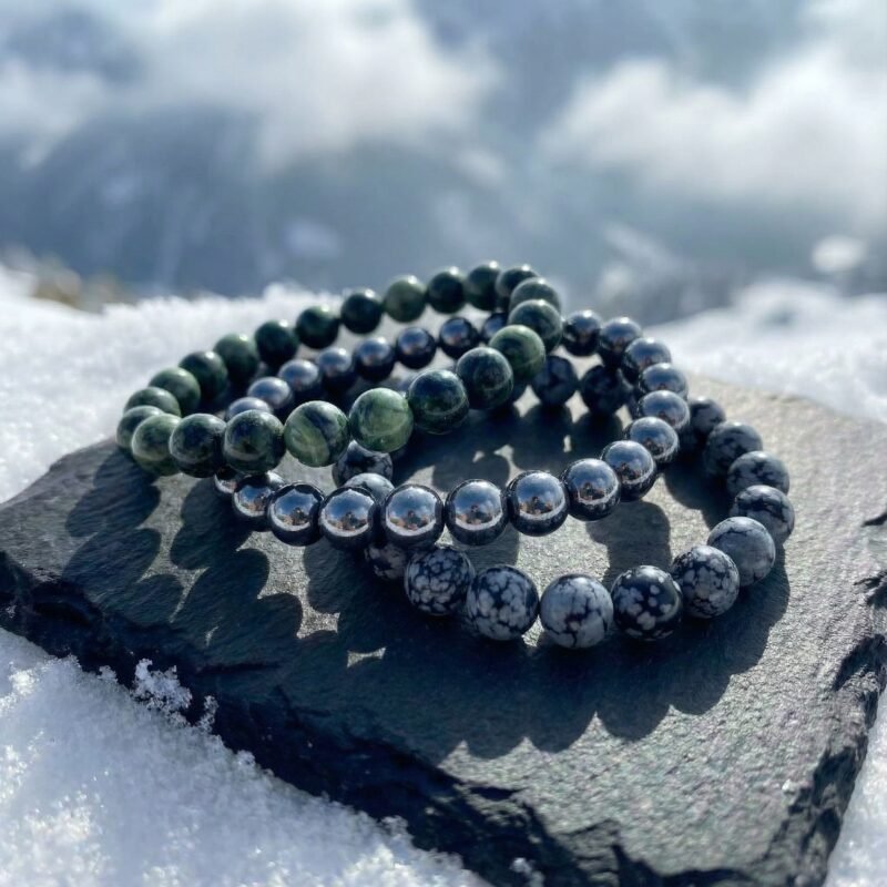 Winter Resilience bracelet triad cascading on a charcoal rock surrounded by snow, deep forest greens and frosted tones inspired by Alpine winter strength.